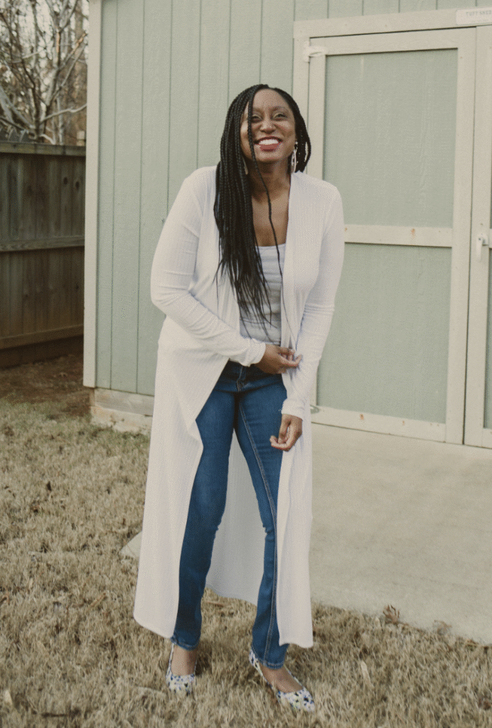 A smiling woman with long braided hair stands outdoors in front of a green shed. She wears a long white cardigan, blue jeans, a gray top, and patterned flats, posing confidently on dry grass.